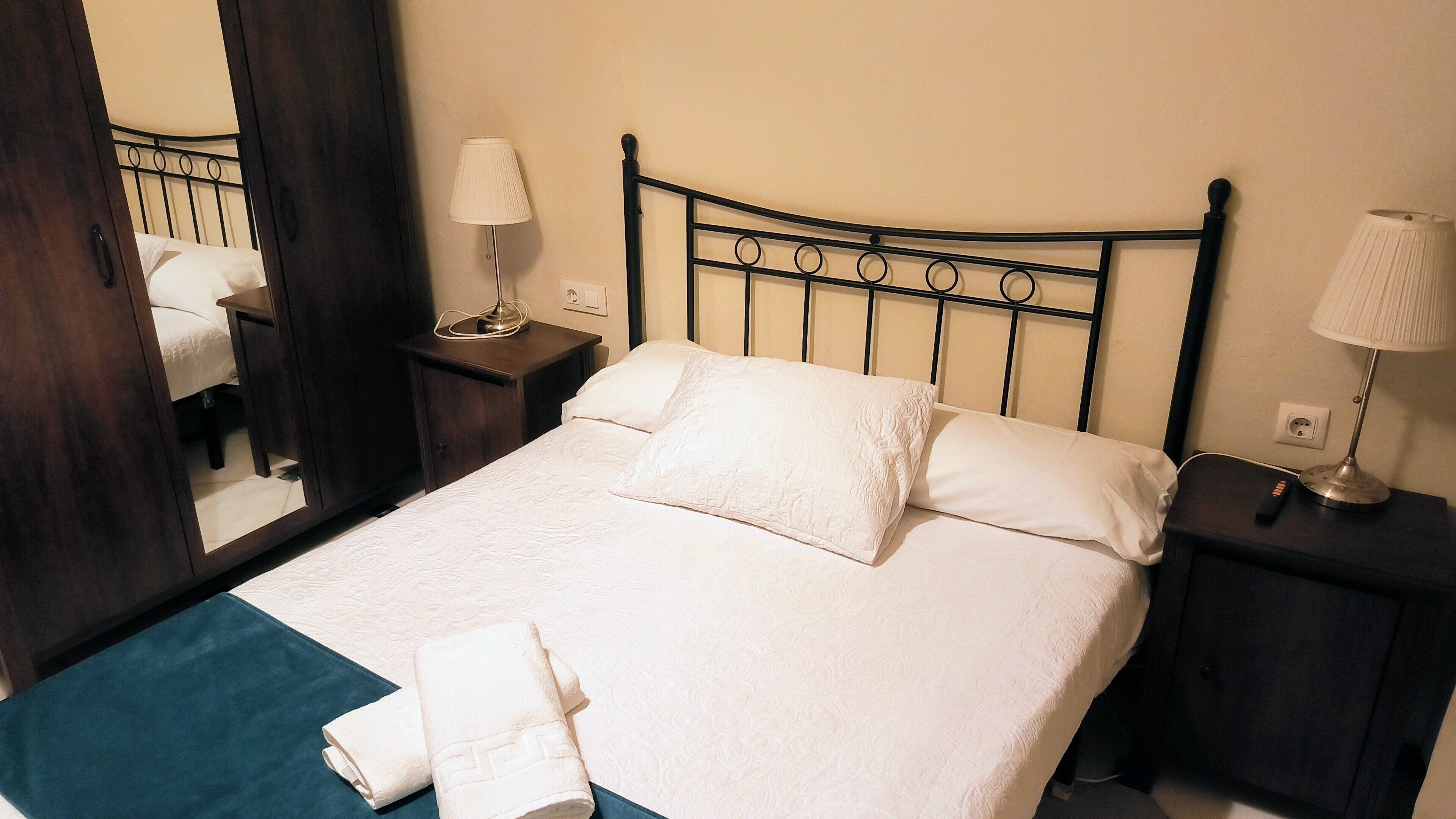 A neatly made bed with white linens and two pillows, a bedside table with a lamp, and a mirror reflecting the room.