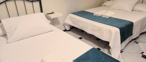A hotel room with two single beds, each with a white bedspread and a teal blanket. There is a nightstand with a remote control and a power outlet on the wall.