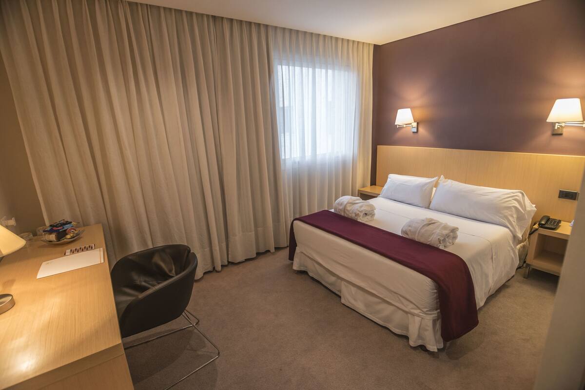 A hotel room with a bed, a desk, a chair, a window with curtains, and two wall-mounted lamps.