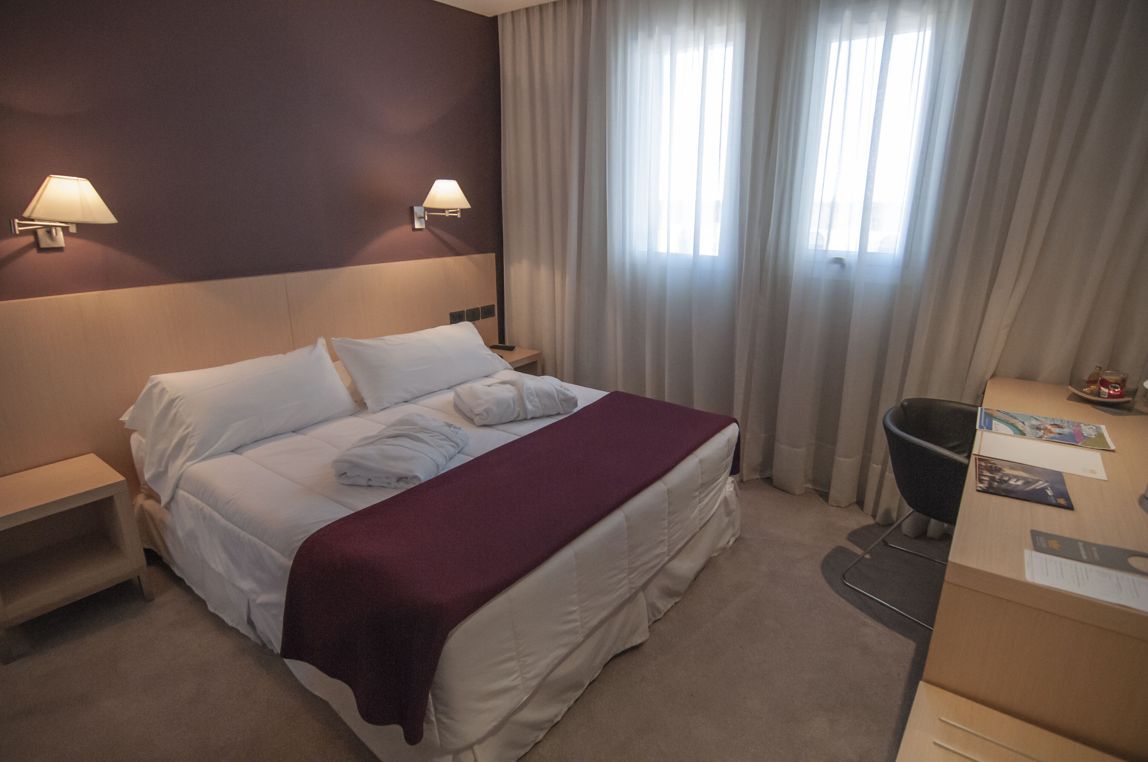 A hotel room with a bed, two bedside lamps, a desk with a chair, and a window with curtains.
