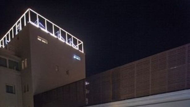 An outdoor swimming pool with a wooden deck, a modern building with a balcony, and a well-lit interior visible through the windows.