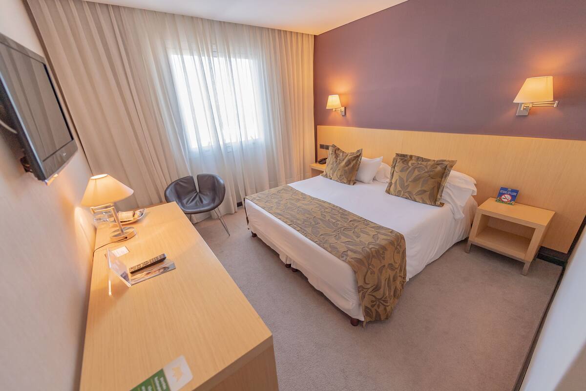 A hotel room with a bed, bedside tables, a desk with a lamp, a chair, and a TV on the wall.