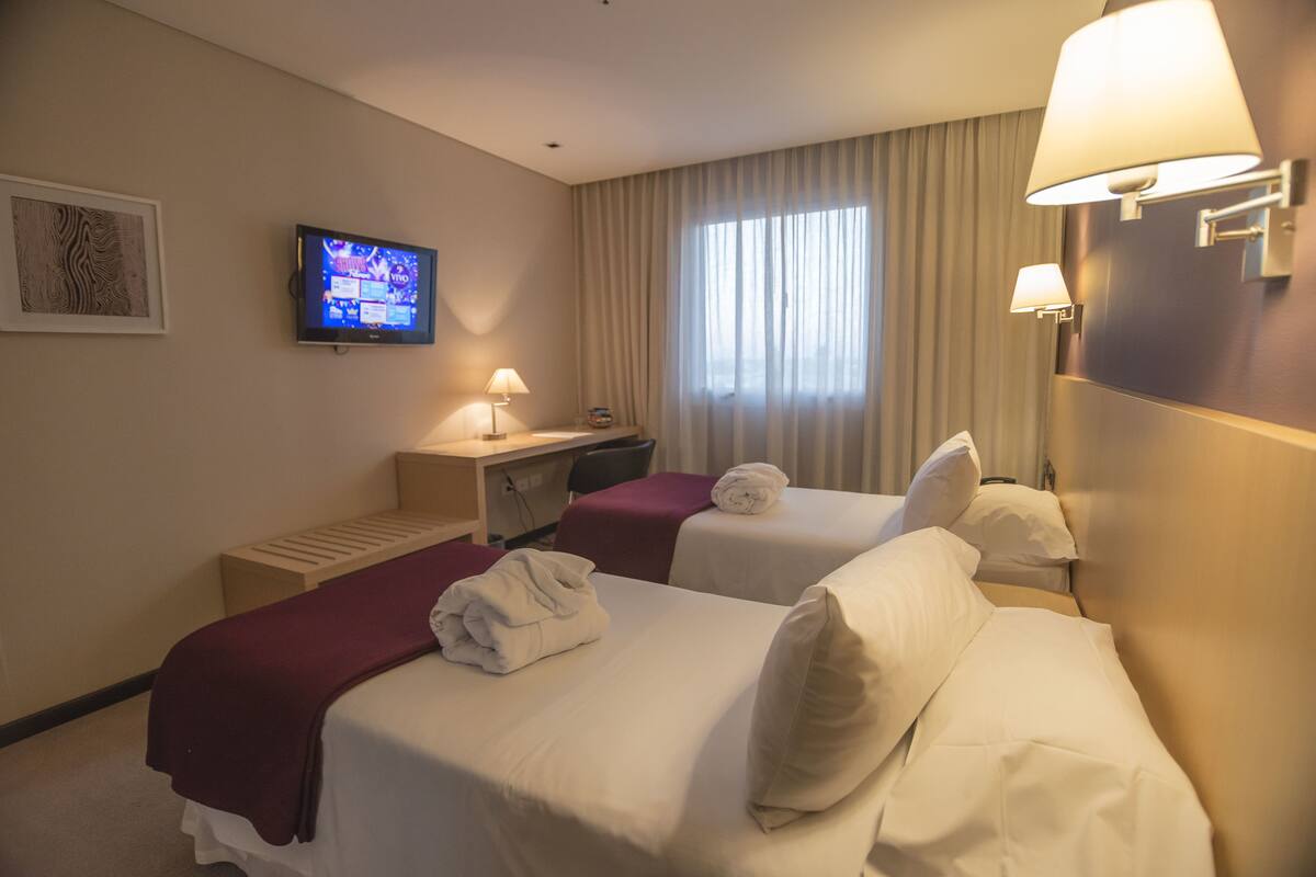 A hotel room with two beds, a desk, a television, and a window with curtains.