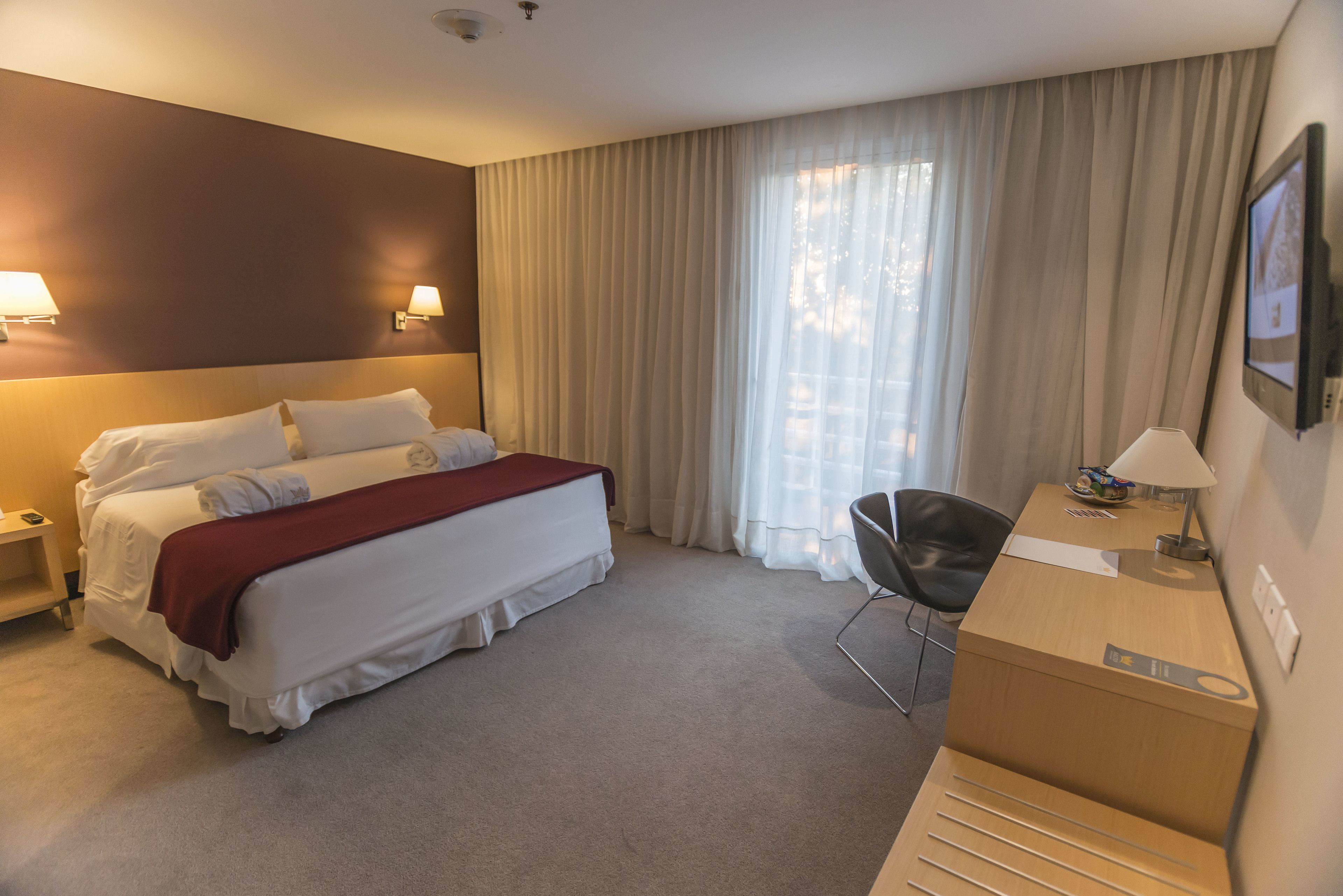 Habitación de hotel con cama, escritorio, silla, lámpara y una ventana grande con cortinas.