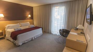 Habitación de hotel con cama, escritorio, silla, lámpara y una ventana grande con cortinas.