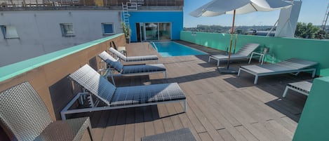 Un área de piscina en la azotea con sillas de descanso y una mesa, un edificio azul al fondo, y un cielo despejado.