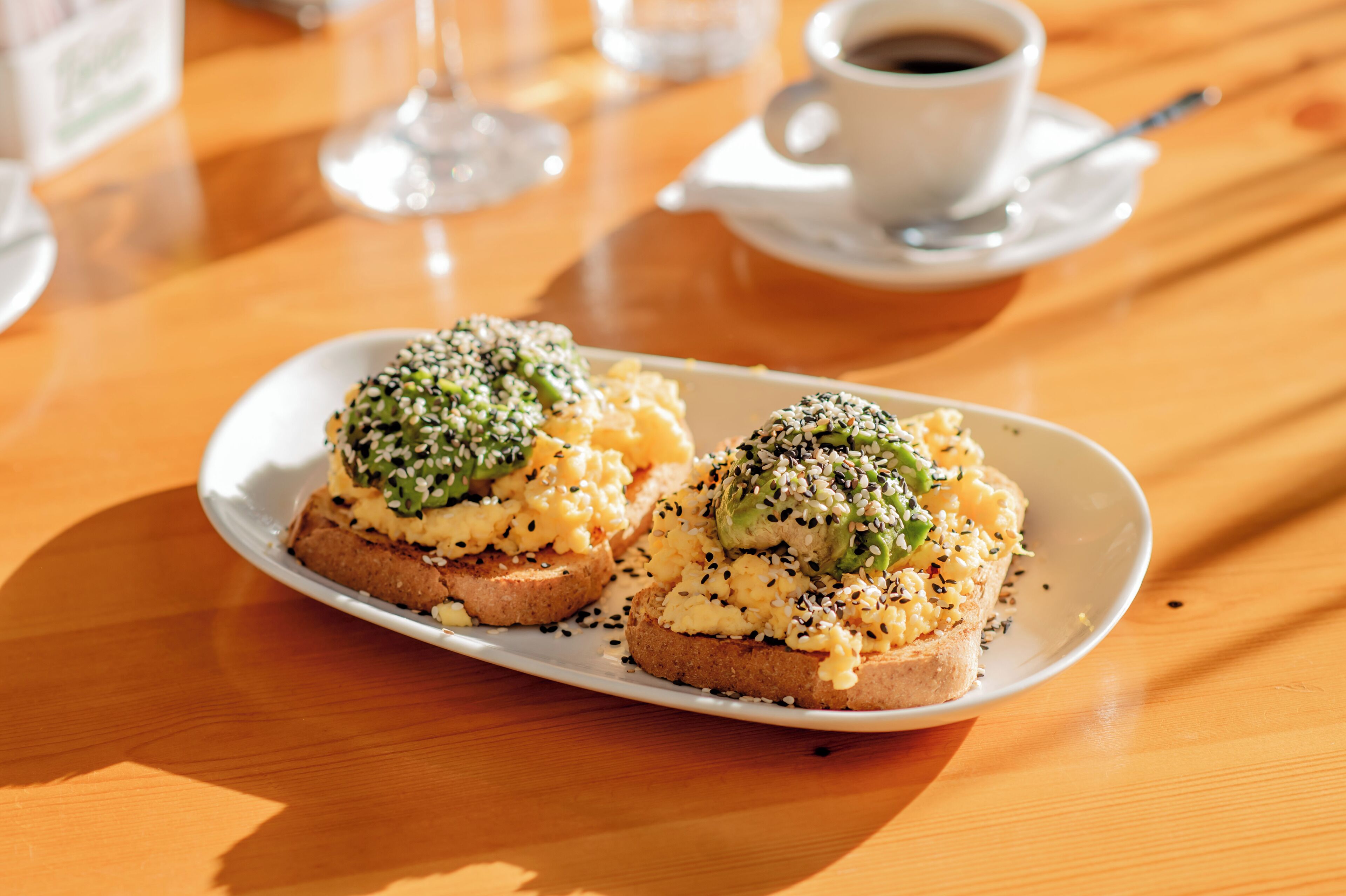 A breakfast plate with scrambled eggs and avocado on toast, a cup of coffee, and a glass of water on a wooden table.