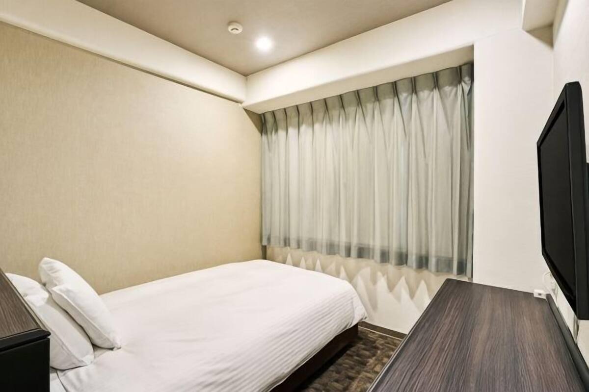 Habitación de hotel con cama, televisión, ventana con cortinas y televisor instalado en la pared.