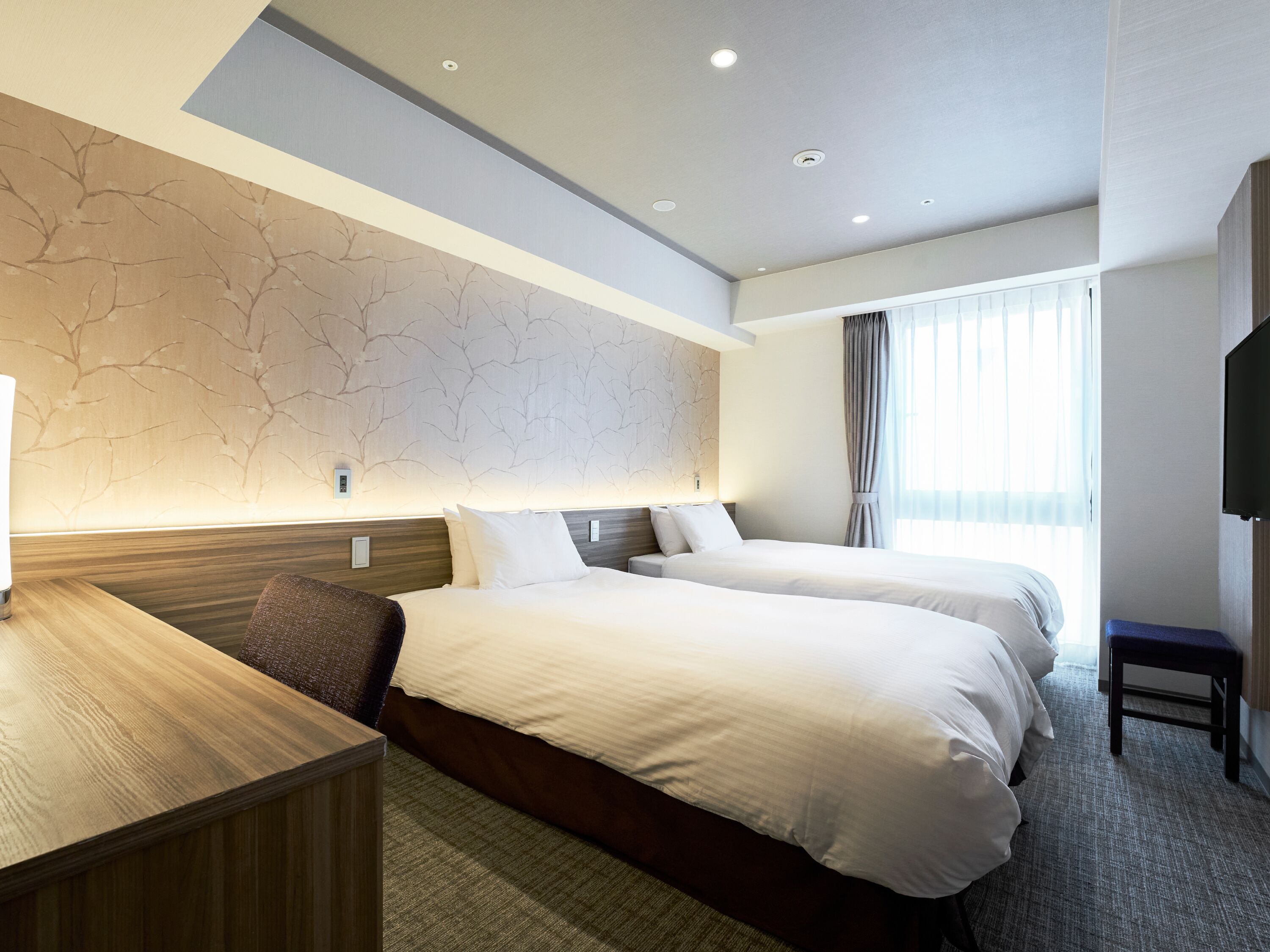 A hotel room with two beds, a desk, a TV, and a large window with curtains.