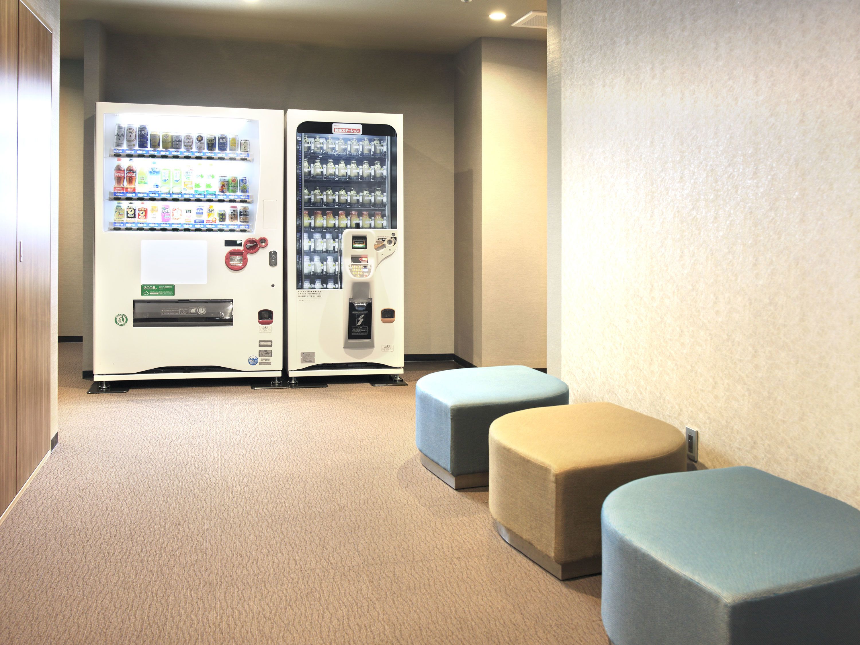 A room with two vending machines, a carpeted floor, and two ottomans.