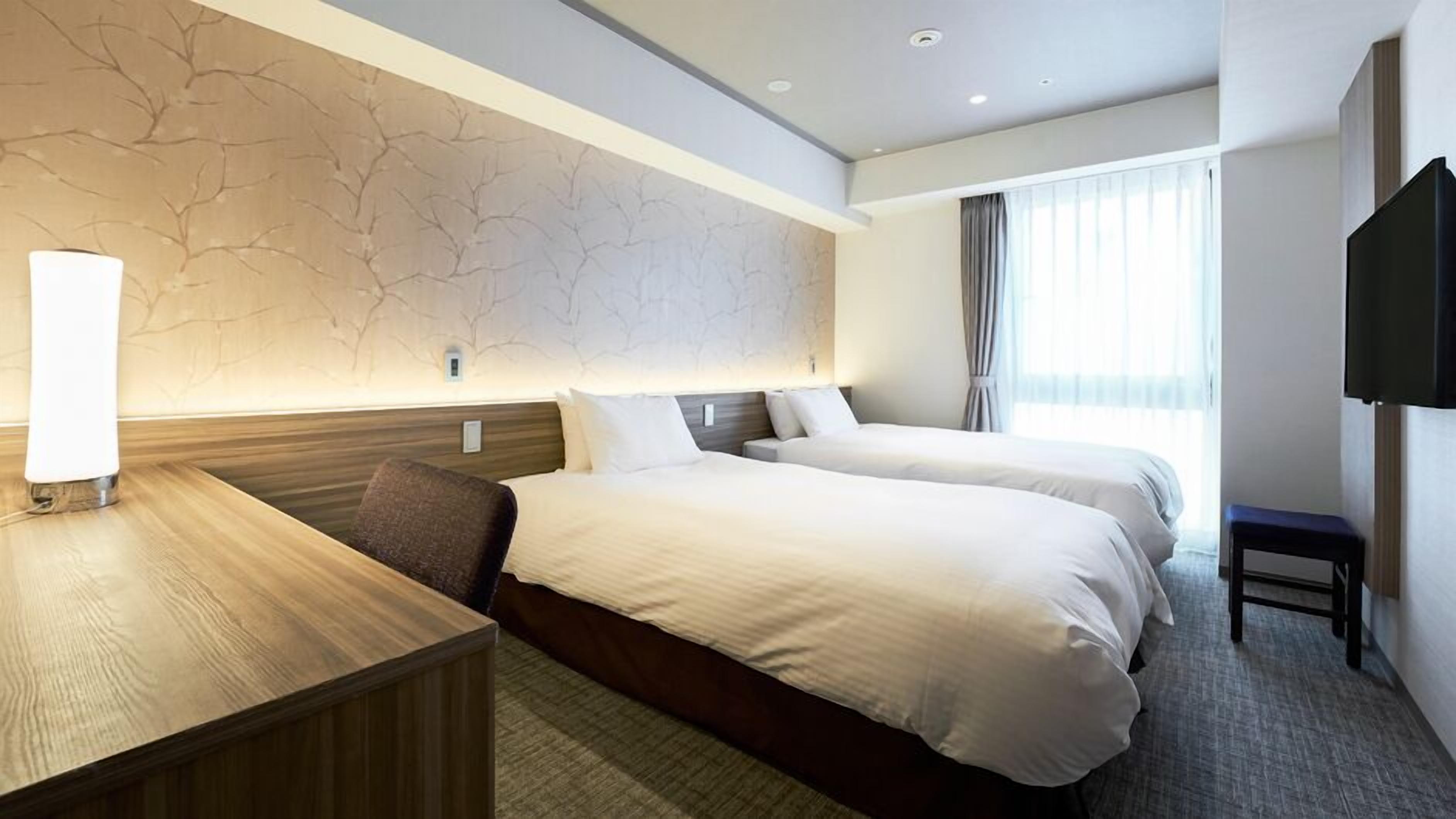 A hotel room with two beds, a desk, a chair, a TV, and a window with curtains.