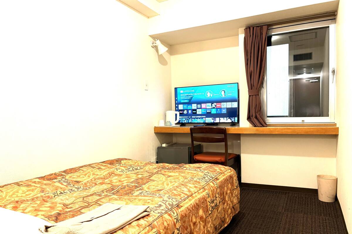 A hotel room with a bed, a desk with a computer, a window, and a small trash bin.