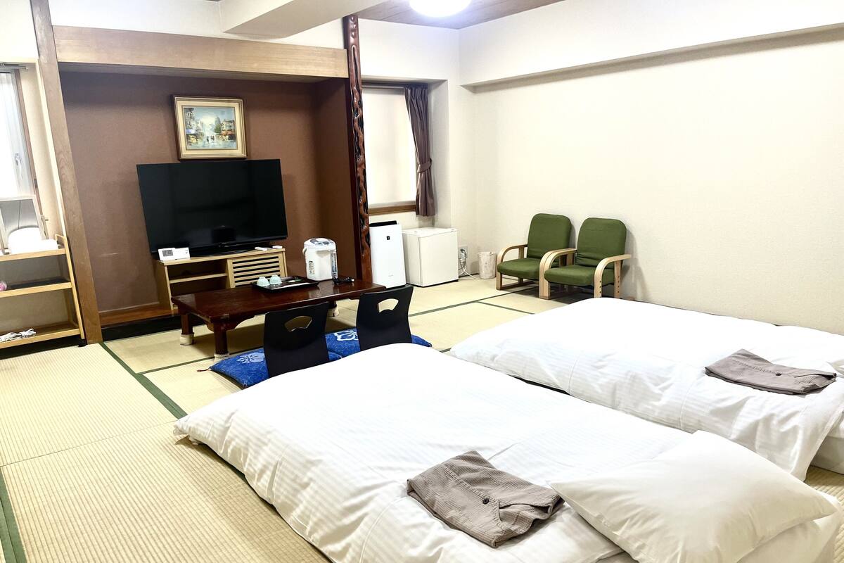 A hotel room with two beds, a television, a desk with a coffee maker, and two green chairs.