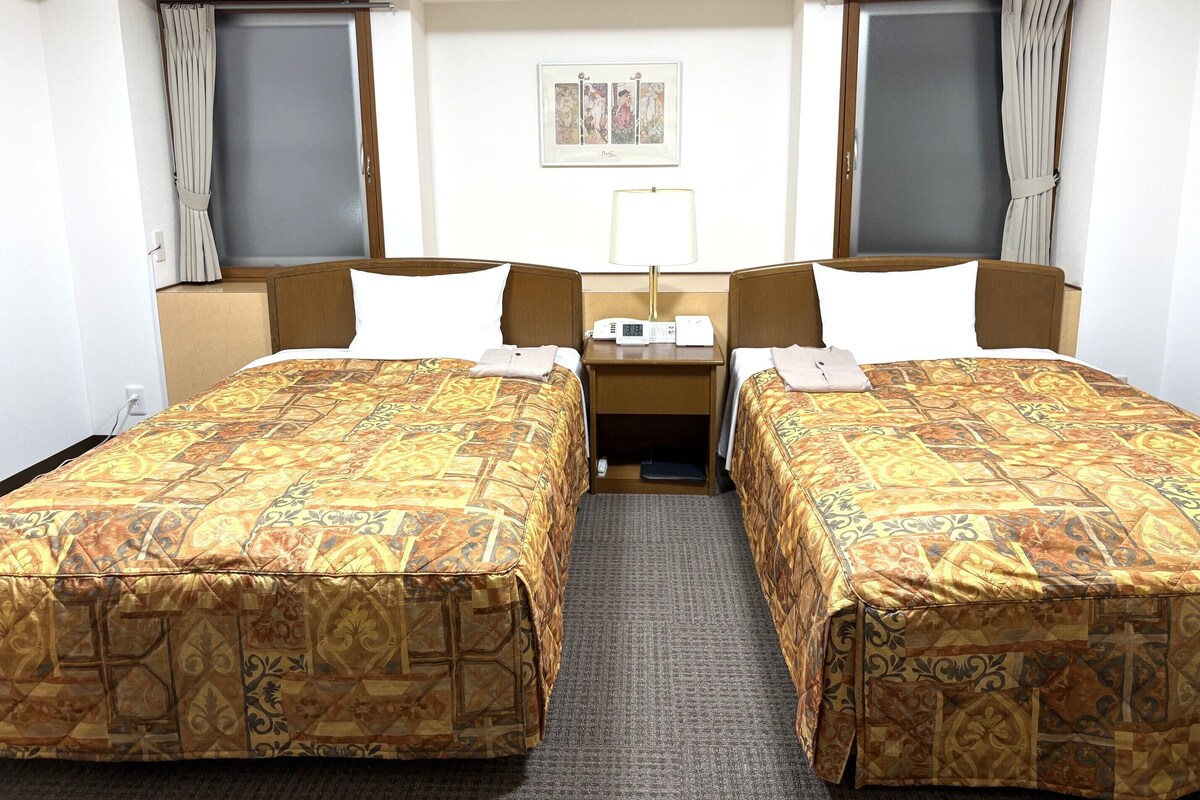 A hotel room with two single beds, a small desk with a lamp, and a framed picture on the wall.