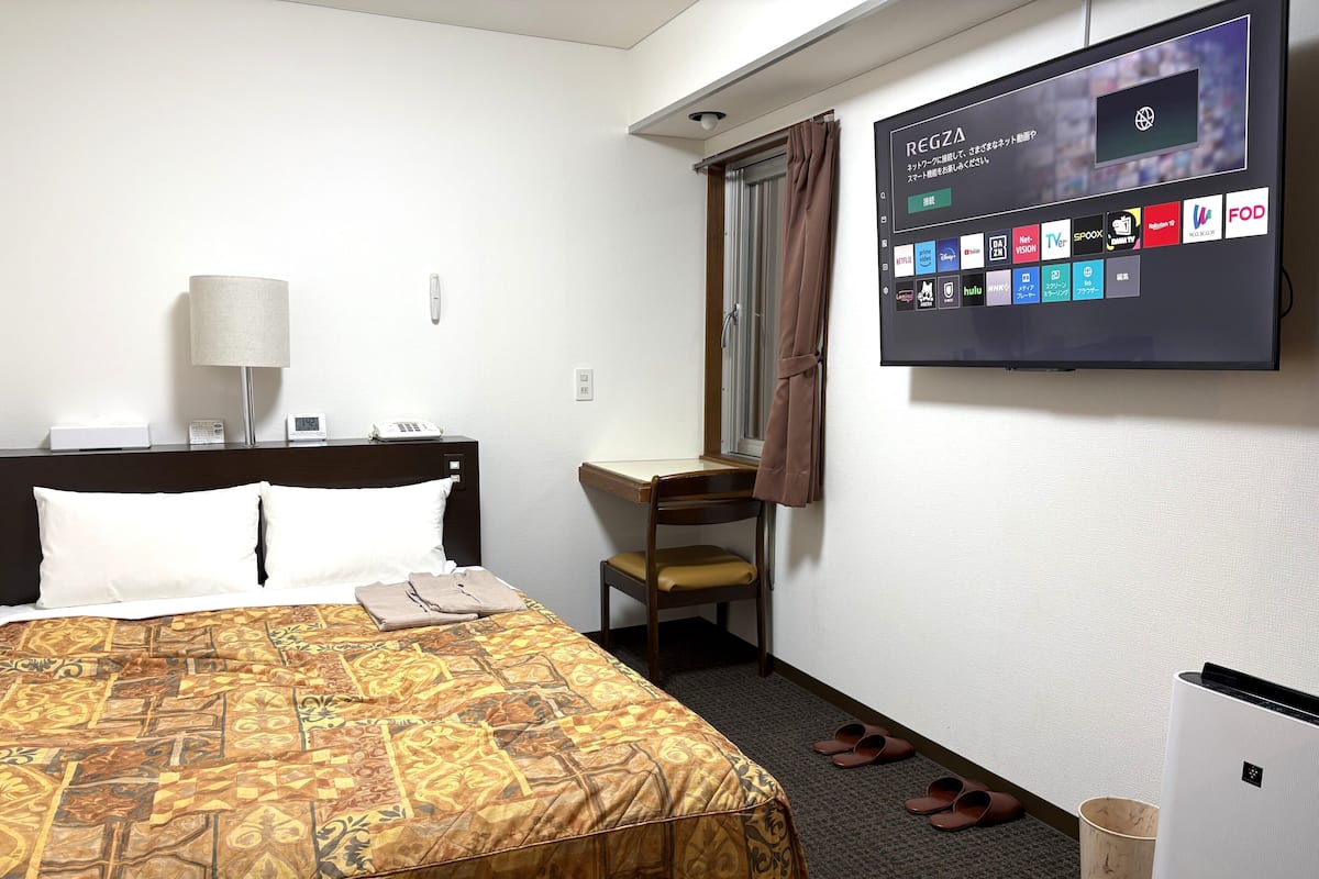 A hotel room with a bed, a desk, a television displaying streaming services, and a pair of slippers on the floor.