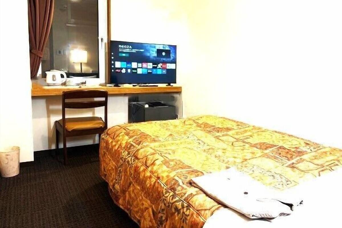 A hotel room with a bed, a desk with a computer, a chair, a window with curtains, and a trash bin.