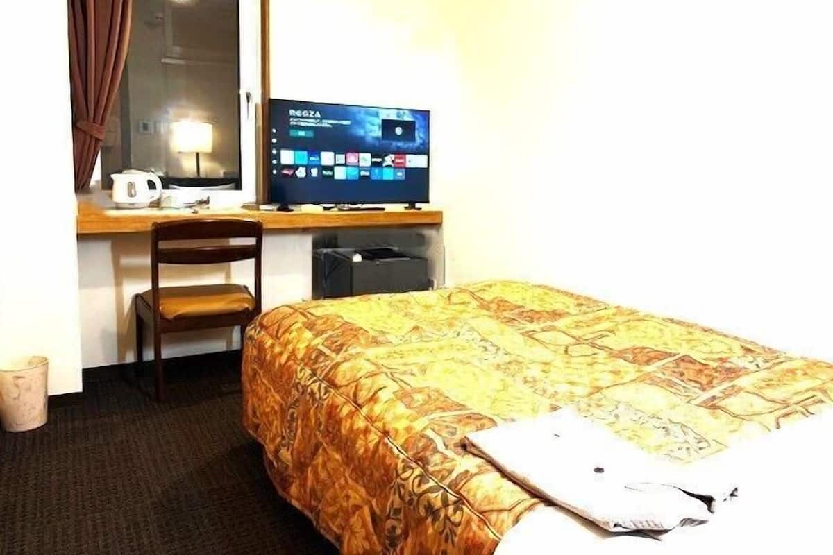 A hotel room with a bed, a desk with a computer, a chair, a window with curtains, and a trash bin.