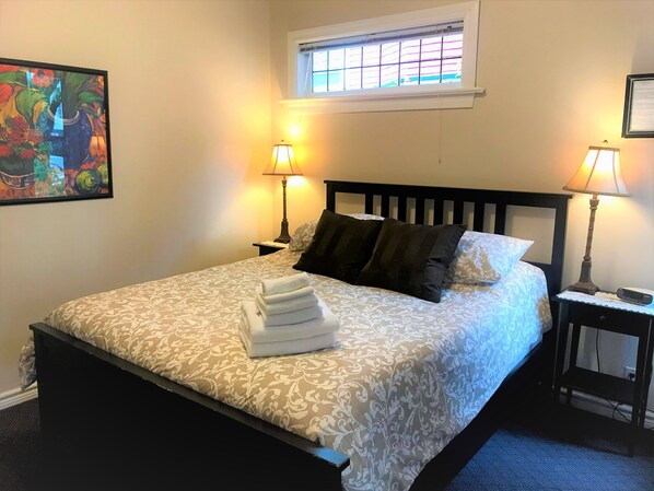 Una cama bien tendida con un edredón estampado, dos lámparas de noche, una ventana con persianas y una obra enmarcada en la pared.