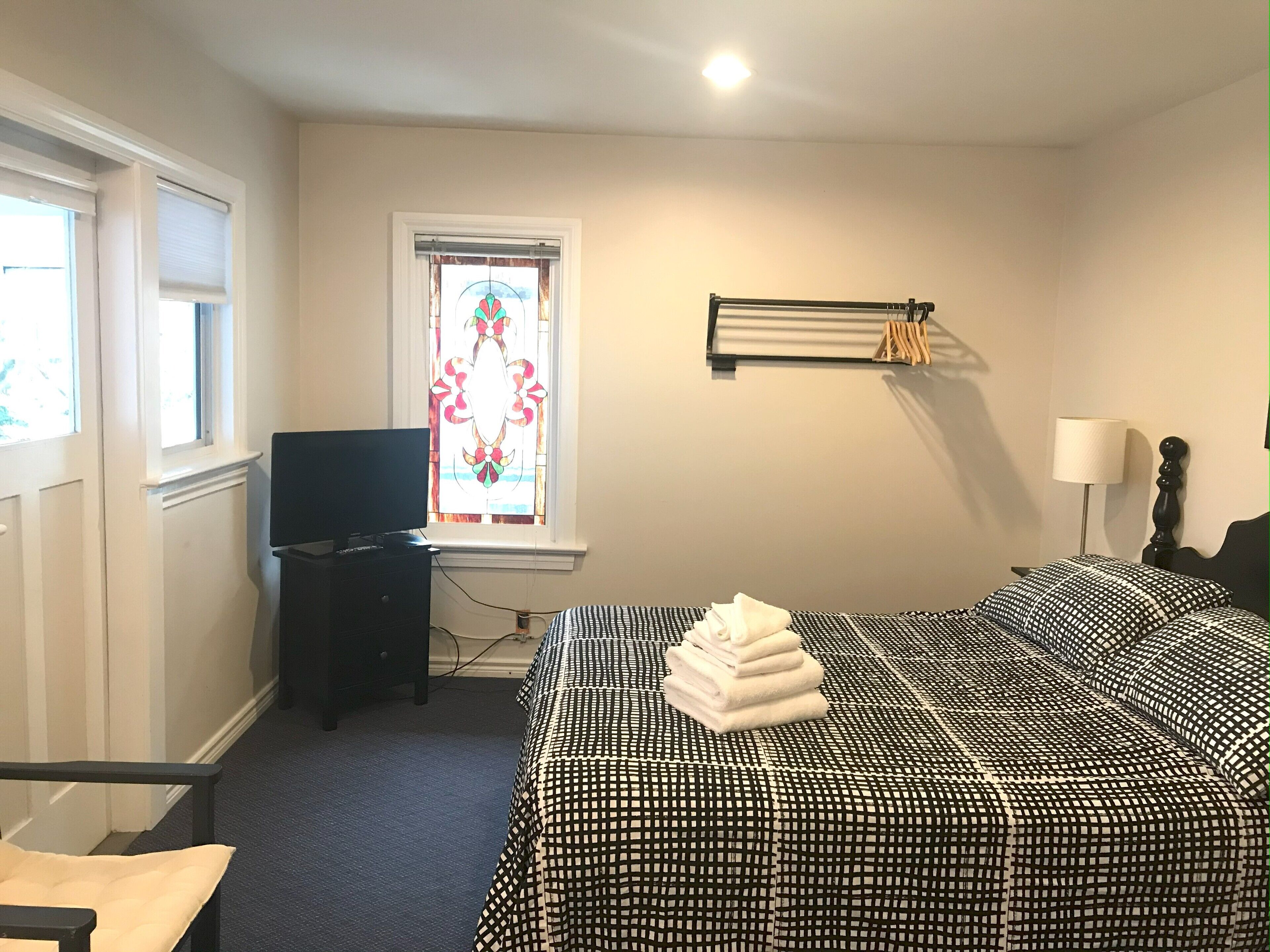 A hotel room with a bed, a television, a window with a stained glass design, a bedside table, and a lamp.