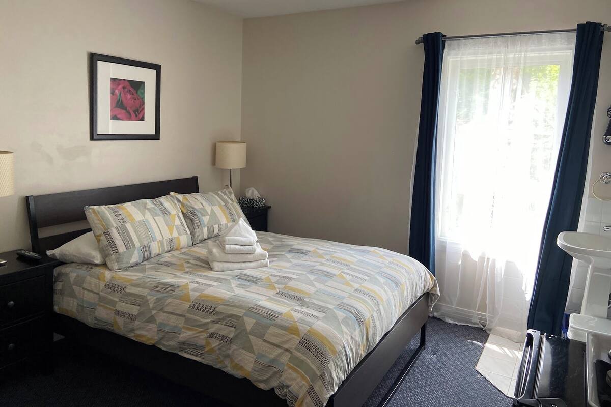 Habitación de hotel con cama, mesita de noche, lámpara, un cuadro en la pared, ventana con cortinas y baño con lavabo y espejo.