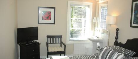 A bedroom with a bed, a chair, a TV, a window, and a bathroom with a sink and mirror.