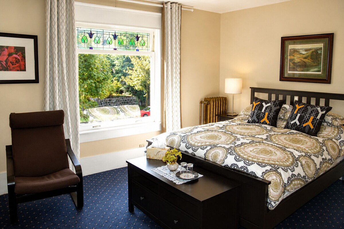 A bedroom with a bed, a chair, a bedside table, a lamp, and a window with curtains.