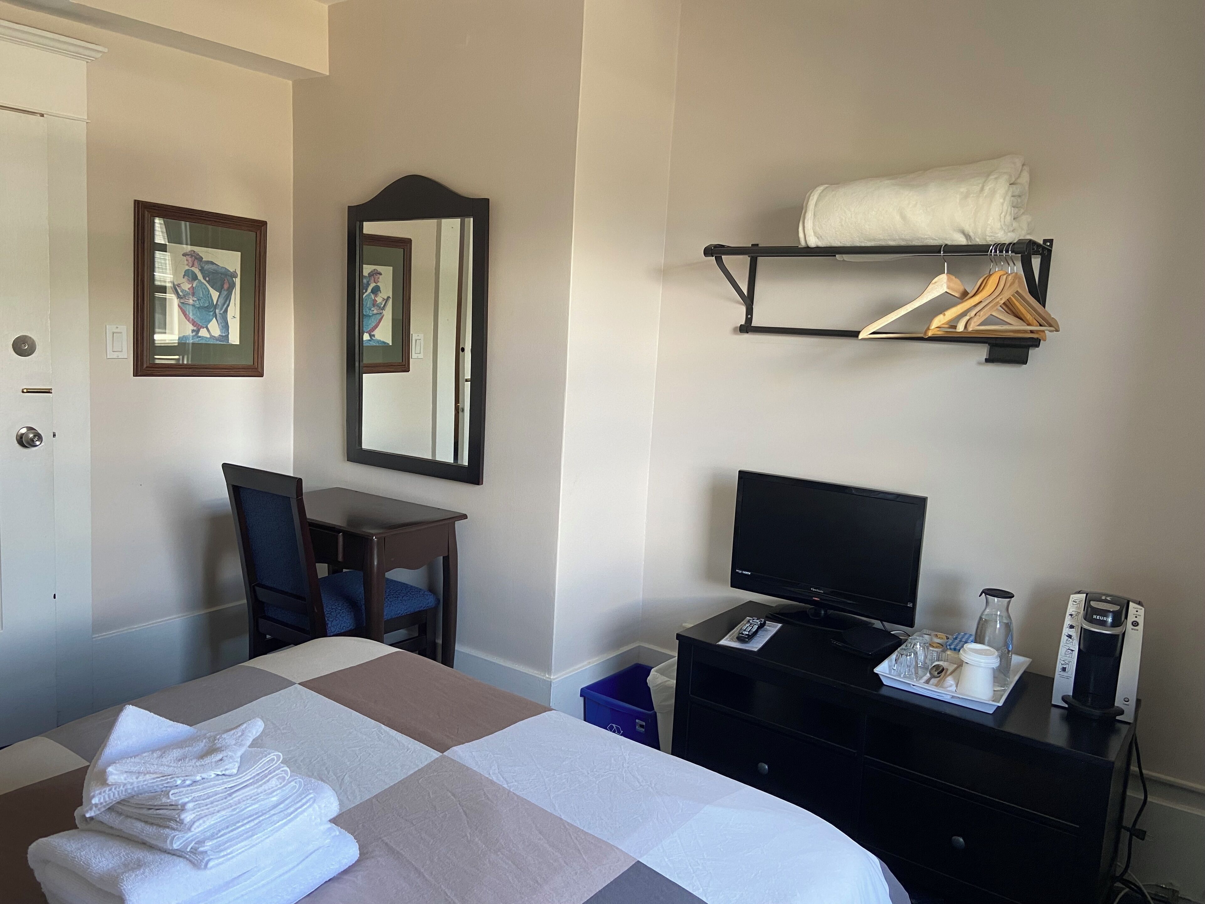 A hotel room with a bed, a desk, a chair, a television, a coffee maker, and a shelf with towels.