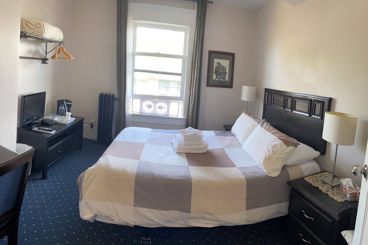A hotel room with a large bed, a desk with a chair, a bedside table with a lamp, a wardrobe, and a window with curtains.