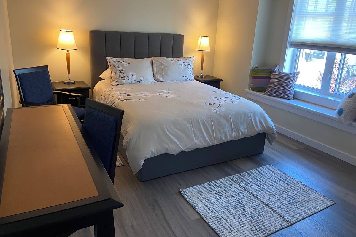 A bedroom with a bed, two bedside tables with lamps, a chair, a desk, a window with blinds, and a view of the outside.