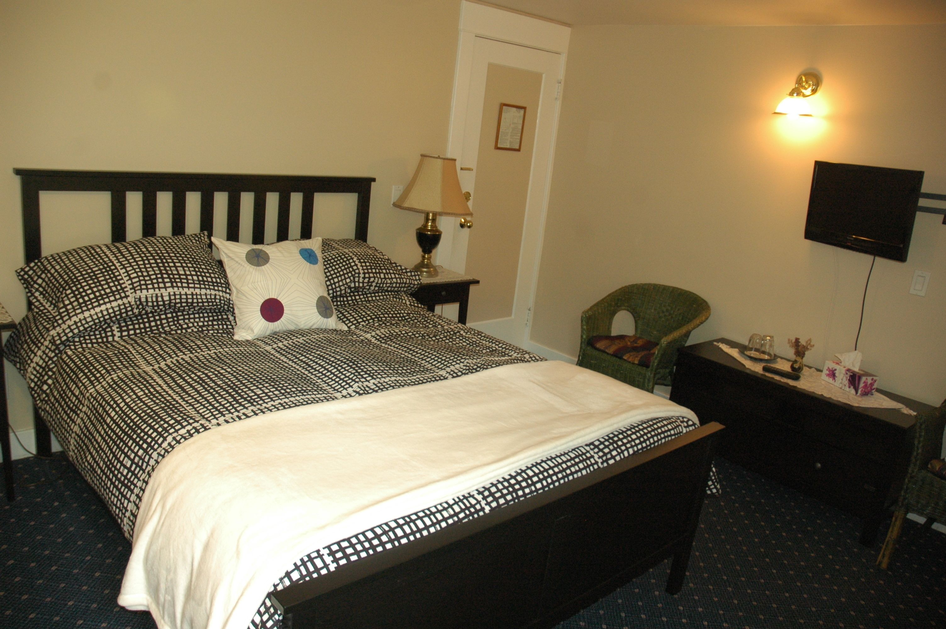 A neatly made bed with patterned bedding, a bedside lamp, a chair, a small table with a lamp, and a TV mounted on the wall.