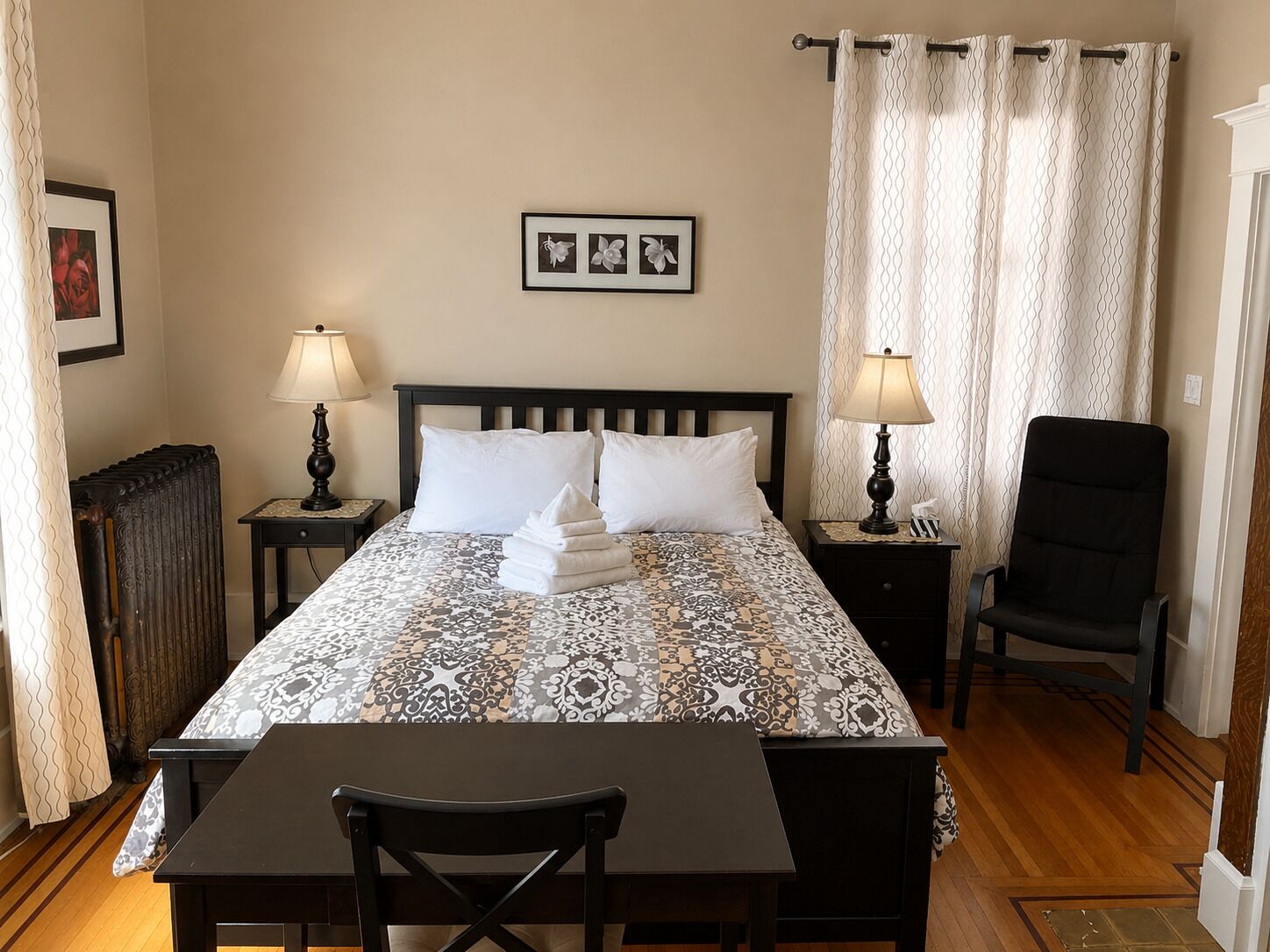 Habitación de hotel con cama, dos mesitas de noche con lámparas, una silla, un radiador y una cortina.