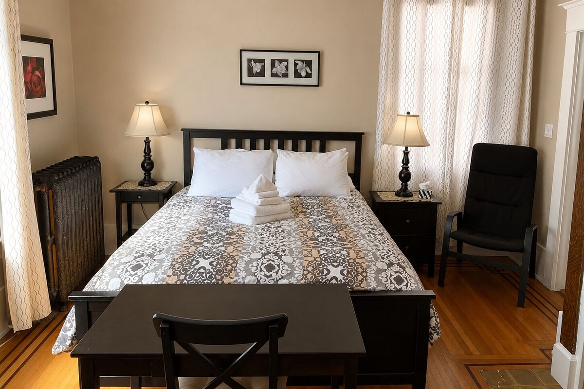 A hotel room with a bed, two bedside tables with lamps, a chair, a radiator, and a curtain.