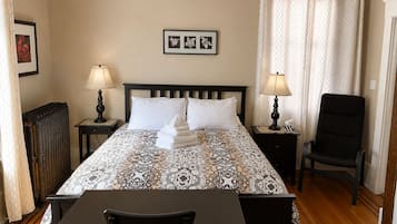 Habitación de hotel con cama, dos mesitas de noche con lámparas, una silla, un radiador y una cortina.