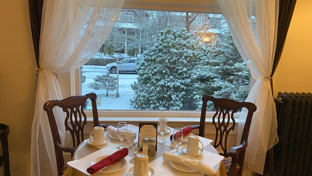 A dining table set with white tableware, a red napkin, and a window view of a snow-covered street. - Windsor Guest House (Vancouver)