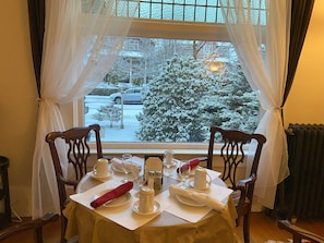A dining table set with white tableware, a red napkin, and a window view of a snow-covered street. - Windsor Guest House (Vancouver)