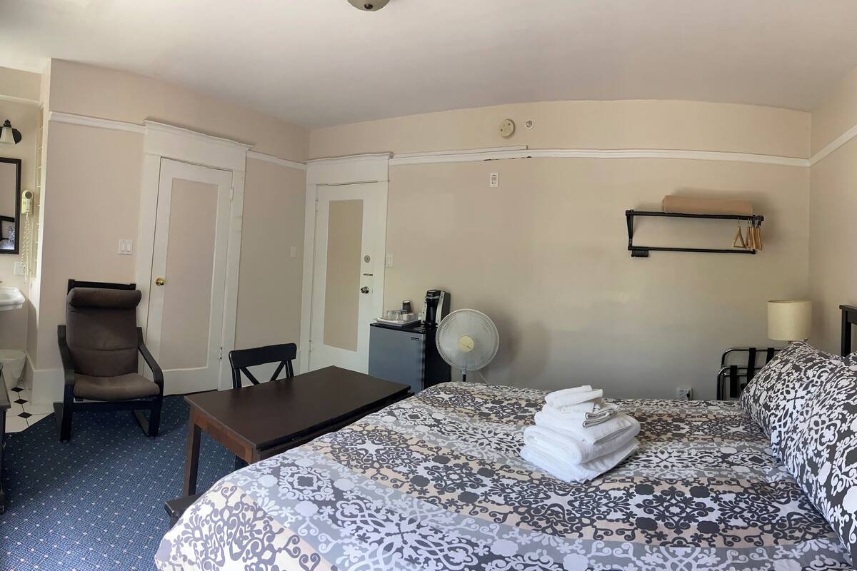 A hotel room with a bed, a desk, a chair, a small table, a fan, and a shelf with towels.