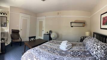 Quarto de hotel com cama, escrivaninha, cadeira, mesa pequena, ventilador e prateleira com toalhas.