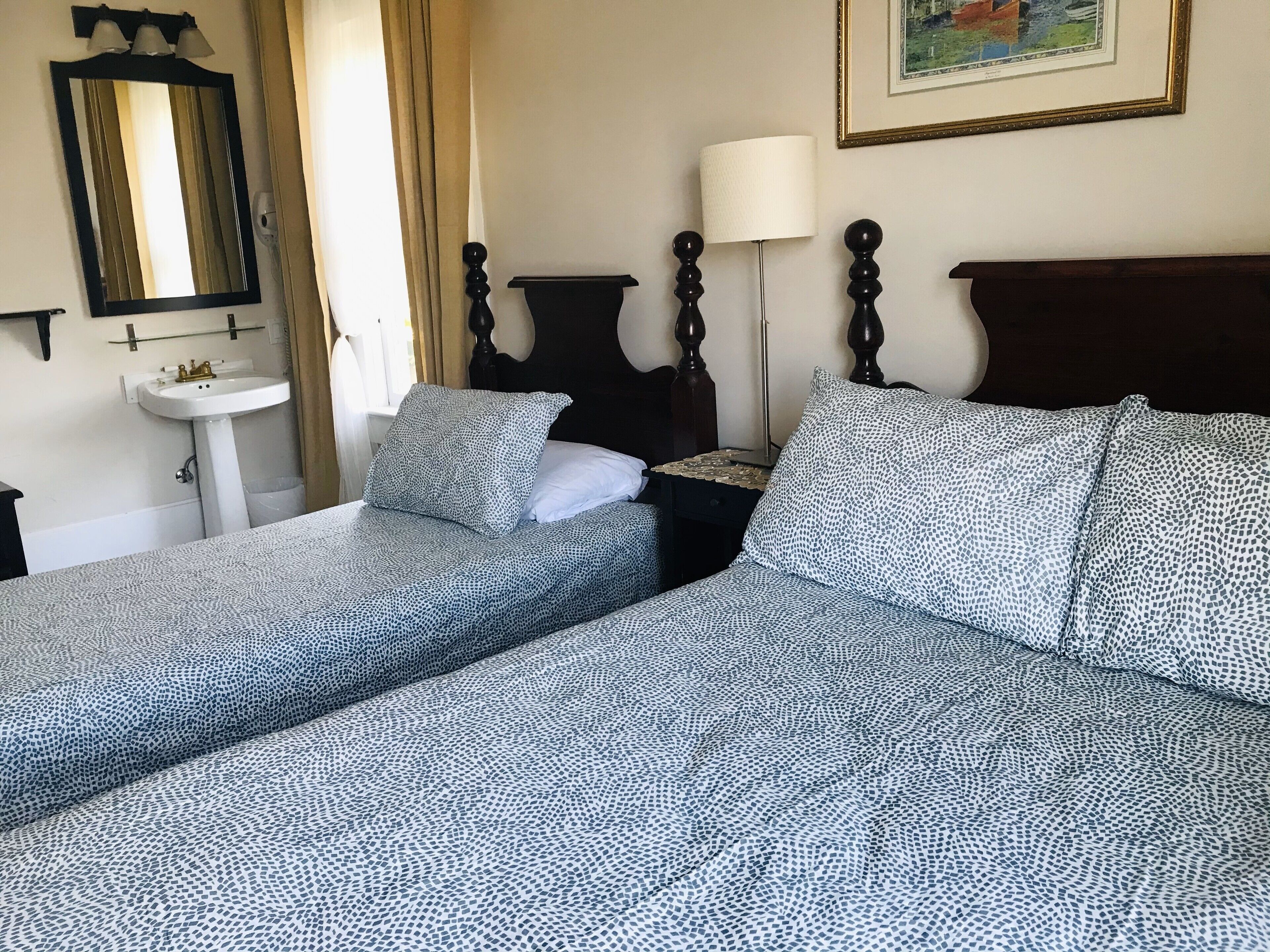 A double bed with a patterned bedspread, a bedside table with a lamp, a nightstand, a mirror, and a wall-mounted shelf.