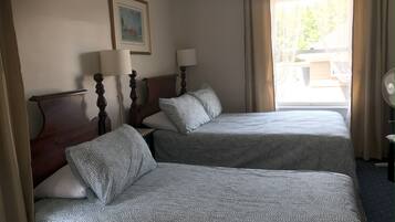 Quarto de hotel com duas camas, cabeceira de madeira, mesa de cabeceira com abajur, um quadro na parede e uma janela com cortinas.
