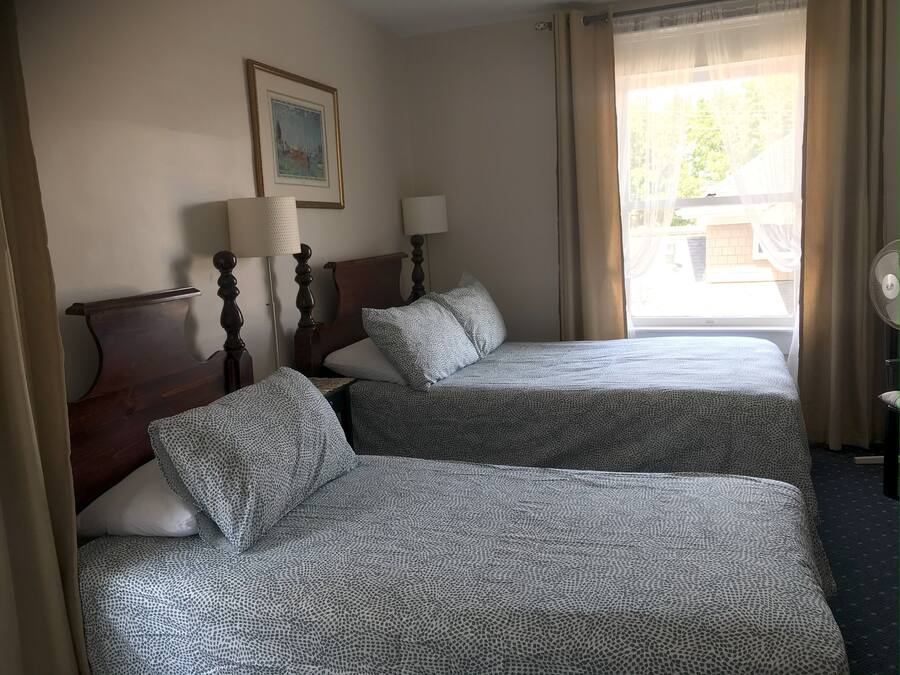 Quarto de hotel com duas camas, cabeceira de madeira, mesa de cabeceira com abajur, um quadro na parede e uma janela com cortinas.