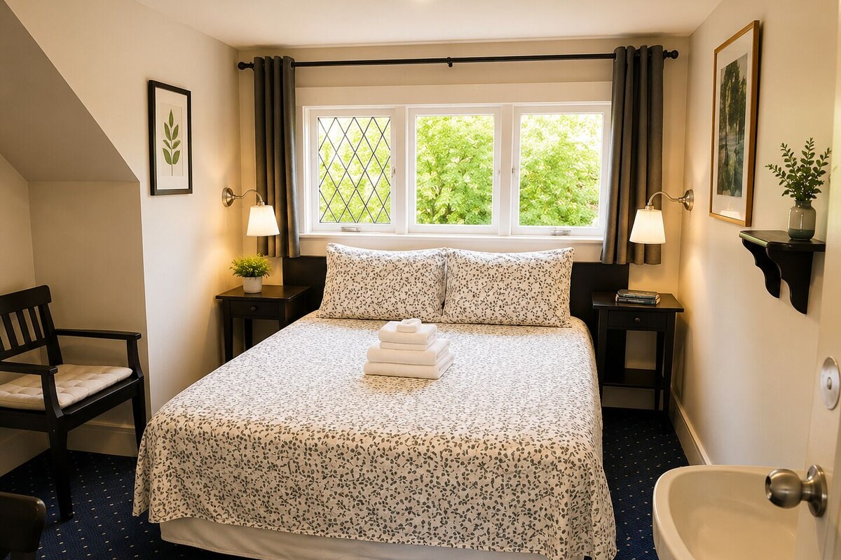 A hotel room with a bed, two bedside tables, a chair, a window with curtains, a potted plant, and a bathroom with a sink and toilet.