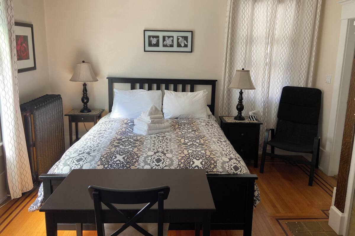 A neatly arranged hotel room with a bed, two bedside tables with lamps, a chair, a desk, and a radiator.