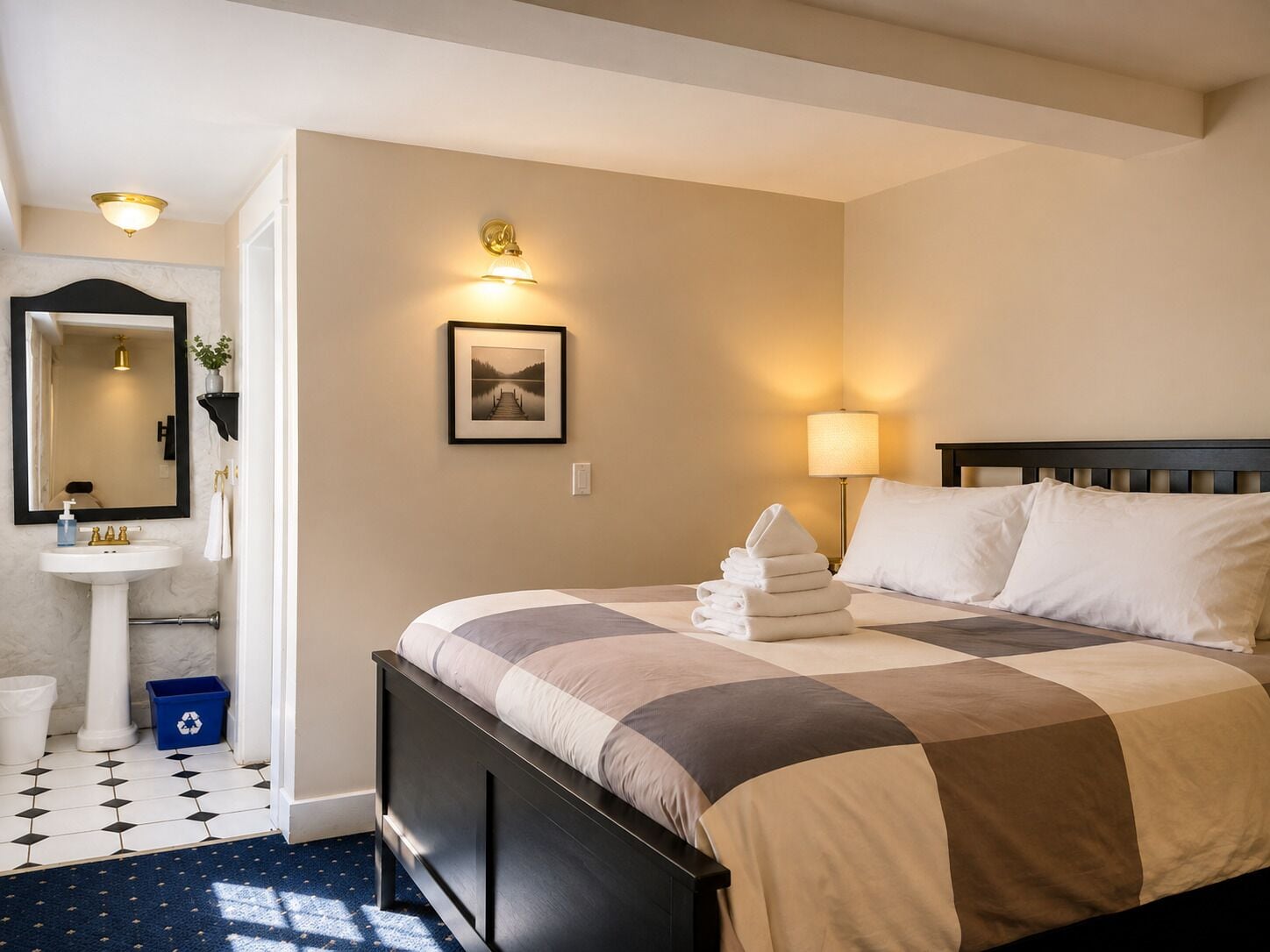 A hotel room with a bed, a bedside lamp, a wall-mounted picture, and a bathroom with a sink and mirror.