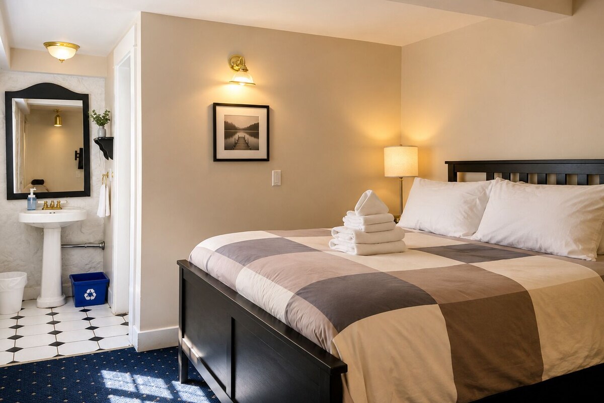 A hotel room with a bed, a bedside lamp, a wall-mounted picture, and a bathroom with a sink and mirror.