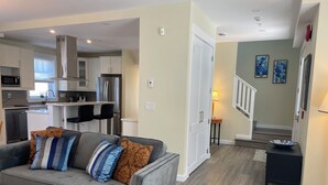 A modern living room with a grey sofa, wooden coffee table, and a dining area with a black table and chairs. A kitchen with white cabinets and stainless steel appliances is visible in the background. - Windsor Guest House (Vancouver)