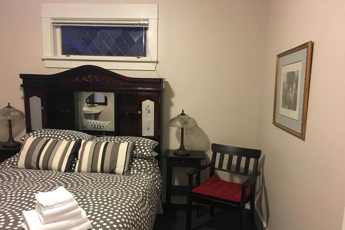 A neatly made bed with striped and polka dot bedding, a chair with a red cushion, a bedside table with a lamp, and a framed picture on the wall.