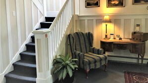 A staircase with white railings and black carpeted steps, a patterned armchair, a desk with a lamp, and framed pictures on the wall. - Douglas Guest House (Vancouver)