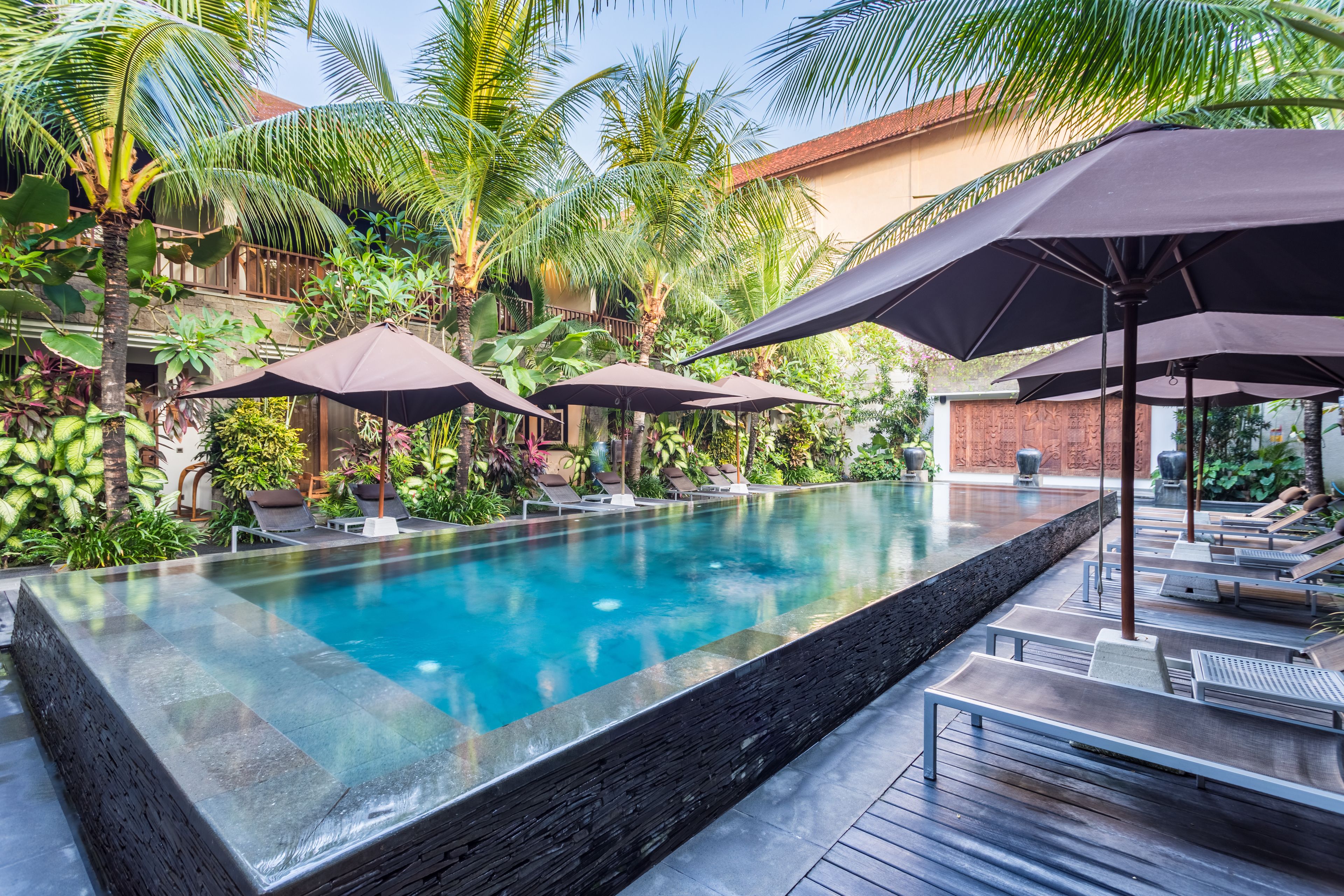 Una piscina infinita rectangular con borde de piedra, rodeada de sillas de descanso y sombrillas, con un fondo de exuberante vegetación y un edificio con tejado de baldosas.