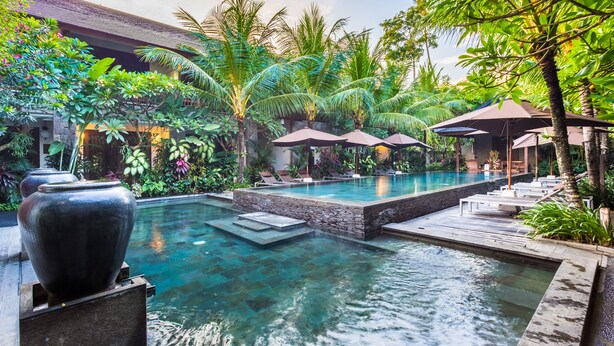 A pool area with lounge chairs and umbrellas, surrounded by lush greenery and a stone wall. - Kejora Suites (Denpasar)