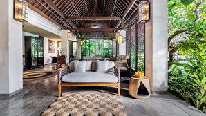 A spacious room with a wooden ceiling, a sofa, a round woven rug, and a view of lush greenery outside. - Kejora Suites (Denpasar)